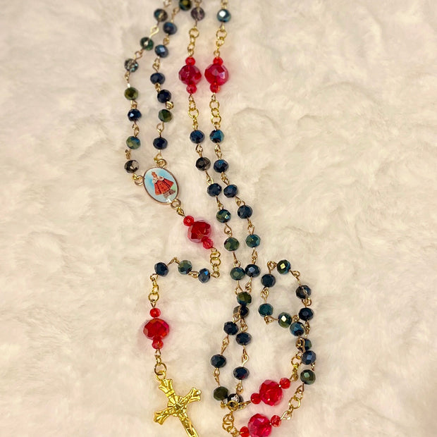 Handcrafted 5-decade dark glass rosary with red crystal Our Father beads, a Baby Jesus center medal, and a gold-toned sunburst crucifix.